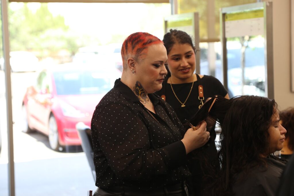 Cosmetology educator Lexie Cochran, with red hair and a black top, holds a section of a guest’s curly hair beside a smiling student artist at a salon station by the front windows.