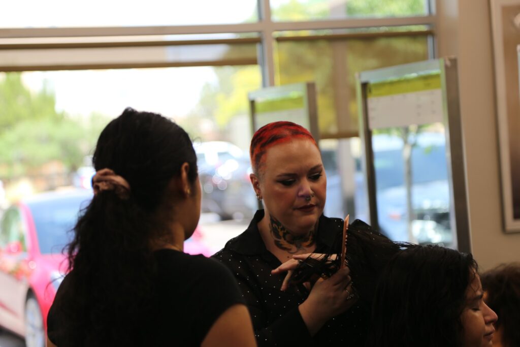 Lexie Cochran talks through a cut at the chair, facing a student artist whose back is to the camera, with a guest seated between them.
