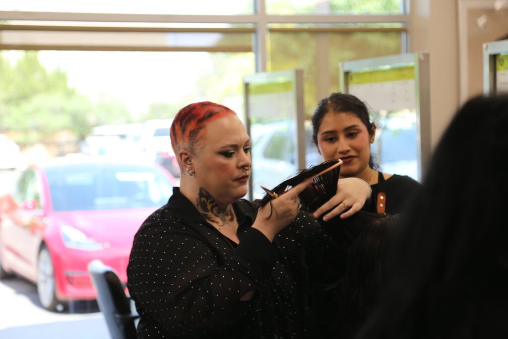 Lexie Cochran holds a comb and shears at a guest’s head while a student artist in an Aveda Institute apron watches closely.