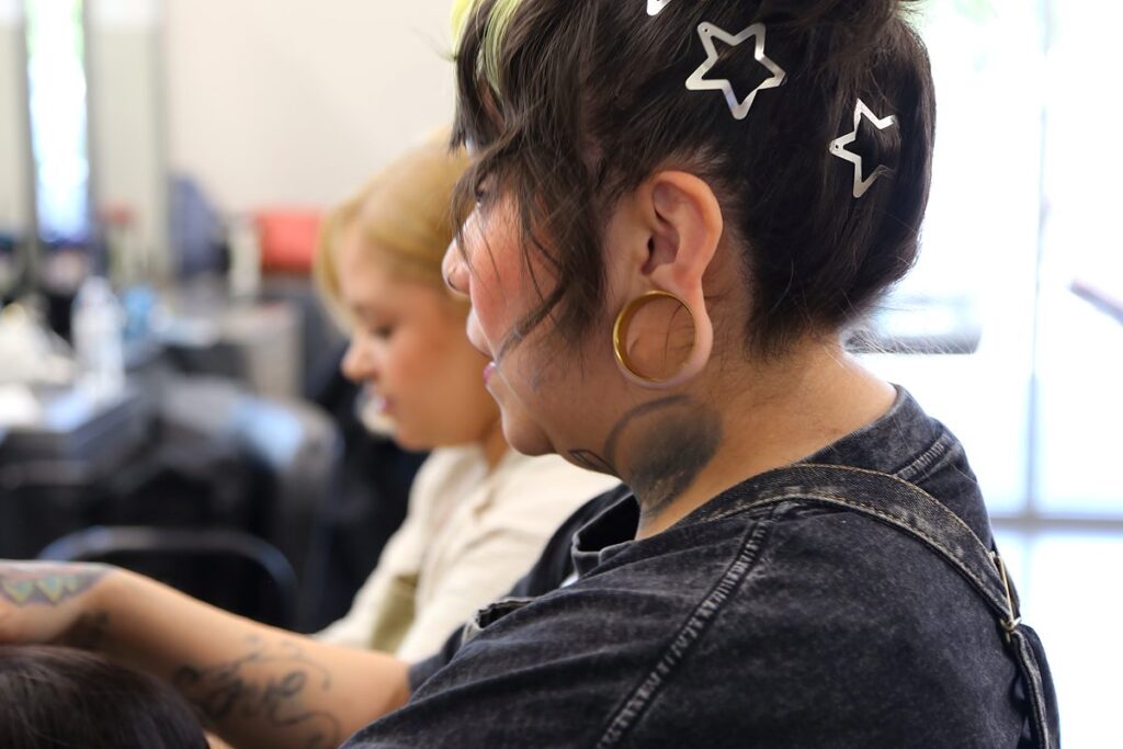 Profile view of cosmetology educator Talia Garcia at a salon station, with a student artist working in the background.