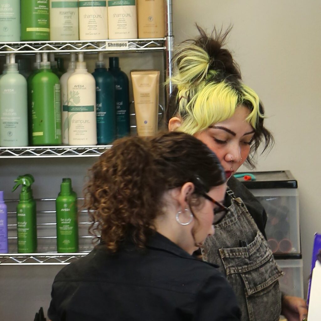 Cosmetology educator Talia Garcia leans in beside a student artist at the dispensary counter, with shelves of Aveda shampoo and conditioner behind them.