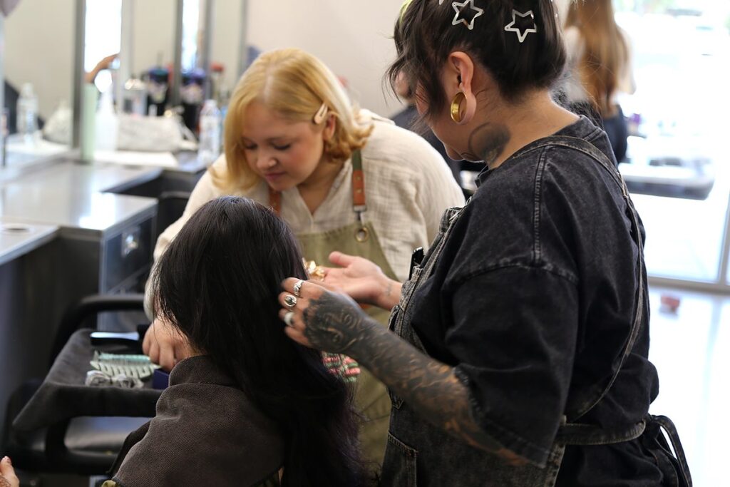 Talia Garcia’s tattooed hands lift a section of a client’s dark hair while a student artist in an apron works on the same client from the opposite side.