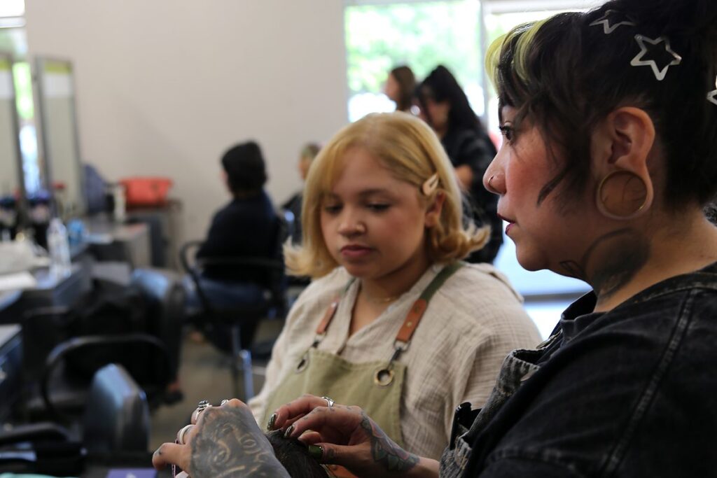 Talia Garcia in denim overalls watches closely over the shoulder of a student artist who is working with her hands at a salon station.